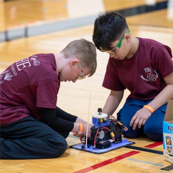 Two children wearing goggles kneel to work on a project on the floor.
