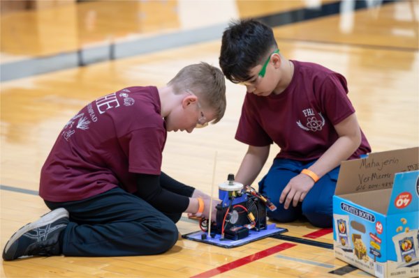 Two children kneel to work on a project that is on the floor.