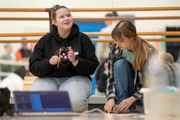 Two students kneel on the floor on a computer. One, holding a picture, smiles while talking. The other student is looking down.