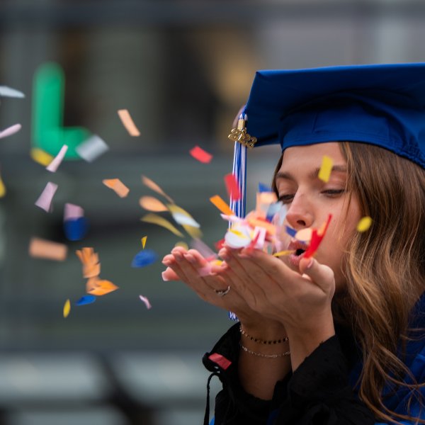 A person wearing a graduation cap with a tassel blows confetti from their hands.