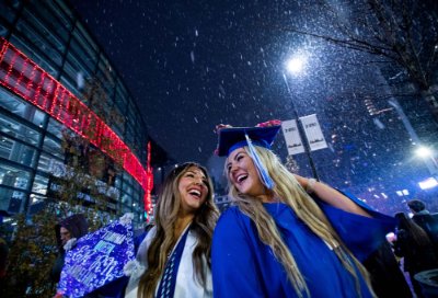 Two people wearing graduation garb smile as they look at each other outside, with snow falling.
