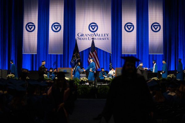 People in academic and graduation garb are on a stage in front of an audience. A banner saying "Grand Valley State University" is in the background.