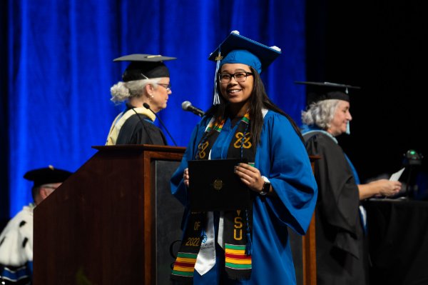 A person wearing a cap and gown and holding a diploma folder looks into the audience.