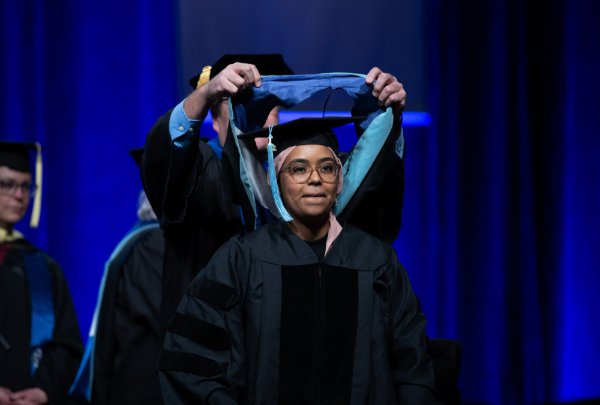 A person wearing graduation garb has an academic hood placed over their shoulders.