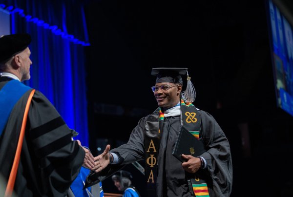 A person wearing a cap and gown and holding a diploma folder shakes the hand of someone in academic garb.