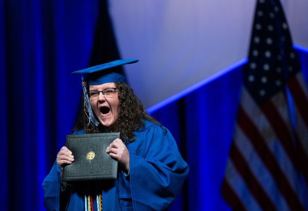 A person wearing graduation garb holds a diploma folder with two hands and gestures with mouth open.