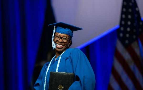 A person wearing a cap and gown and holding a diploma folder smiles broadly on stage.