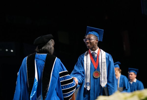 A person wearing graduation garb smiles at a person wearing academic garb while shaking their hand.