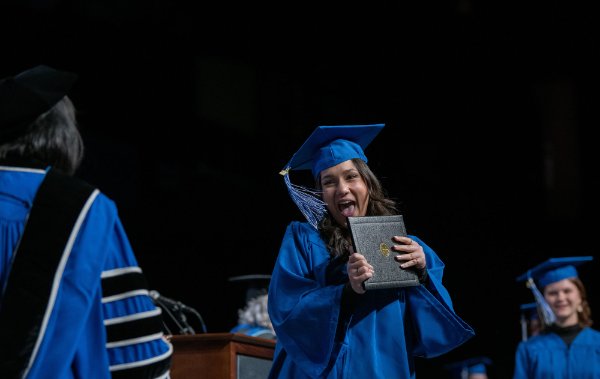 A person wearing a cap and gown and holding a diploma folder smiles broadly.