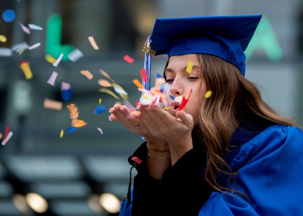A person wearing a cap and gown blows confetti from their hands.