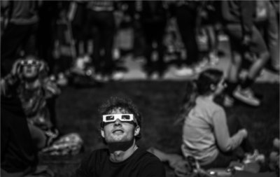 A person wearing special glasses for a solar eclipse looks skyward while sitting on the ground. Other people sit behind the person.