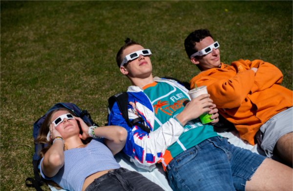 Three people lie on the ground while wearing special glasses for an eclipse. They are looking skyward.