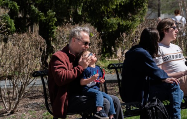 A child sitting on a person's lap looks skyward while the adult helps keep the special eclipse glasses secure.