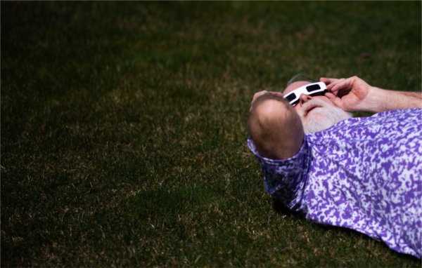 A person lying on the ground holds special glasses against their head while viewing the solar eclipse.