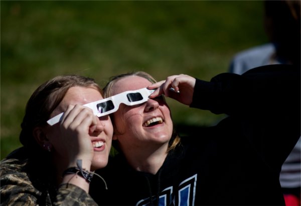 Two people smile while looking upward. They are sharing a pair of special glasses for a solar eclipse.