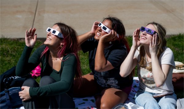 Three people sitting on the ground smile while looking upward and wearing special solar eclipse glasses.