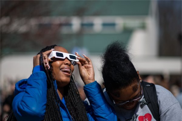 One person wearing special glasses and looking upward smiles while another person looks down while laughing.