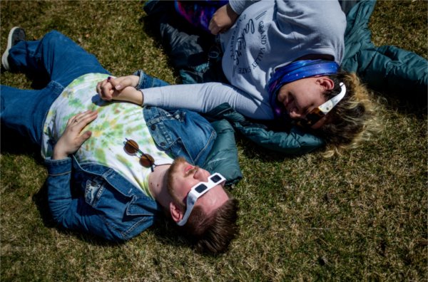 Two people hold hands while lying on the ground and wearing special glasses to view the solar eclipse.