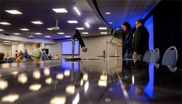  Two people stand at a lectern as they address a crowed as overhead lights reflect onto a shiny table. 