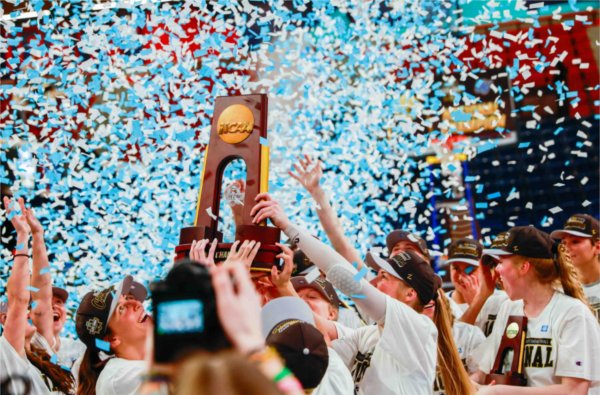 Colorful blue and white confetti falls from an arena as a college women's basketball team hoists a national championship trophy into the air as they celebrate a win. 