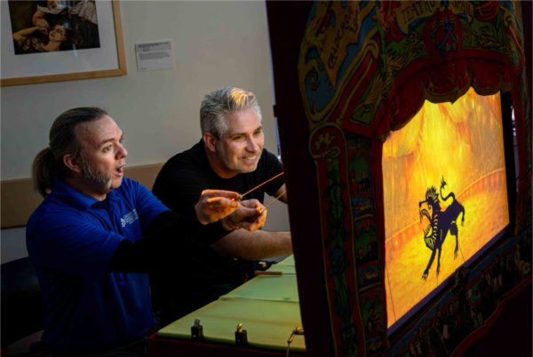  Two people make expressive faces as they work puppets behind the scenes of a puppet theatre. 