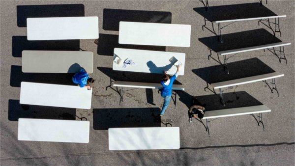   As seen from a drone, students wearing blue clean off white folding tables in a row. 