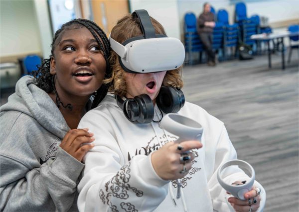  Two students make expressive faces as one uses a VR headset. 