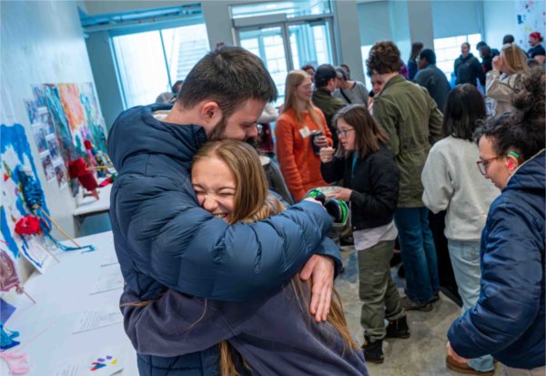 Two people embrace with one laughing during an art event. 