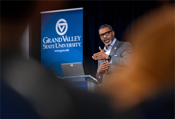   A man gestures as he speaks near a blue Grand Valley State University banner. 