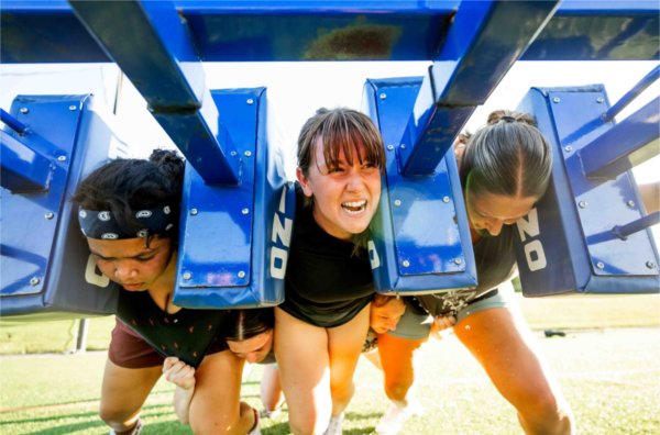 Women rugby players grimace as they push a blue sled. 