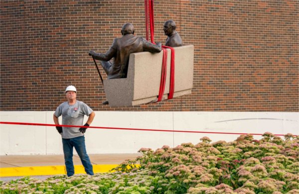 A person wearing a hard hat oversees a sculpture as it is lowered into place by red straps. 