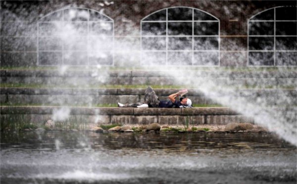  A college student wearing a bandana lays on a ledge reading a book by a fountain. 