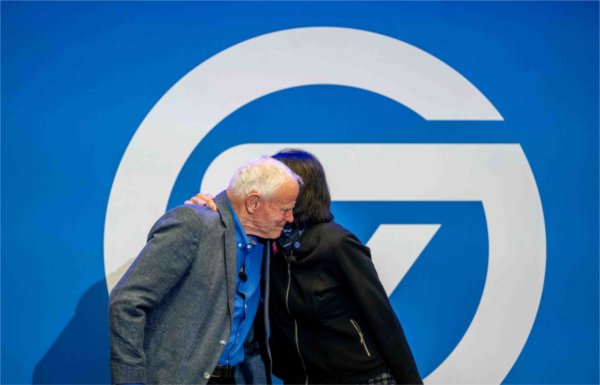 Two people embrace in front of a large college logo in blue and white. 