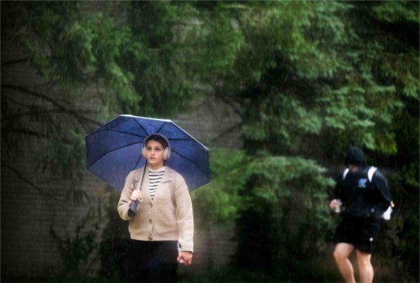  A college student wearing headphones and carrying an umbrella walks through the rain. 