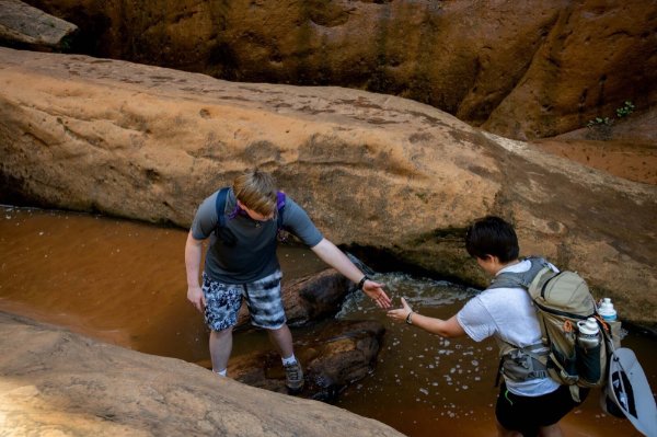 A person reaches back to give a hand to another person as they walk through a crevice between rocks.