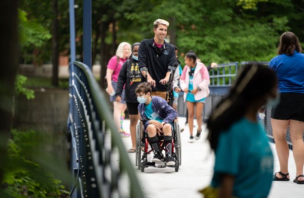  A person pushes another person in a wheelchair across a blue bridge surrounded by several others walking by.