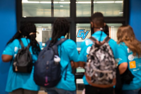 The backs of four people wearing backpacks look through a window toward a "GV" logo on the wall in the background.