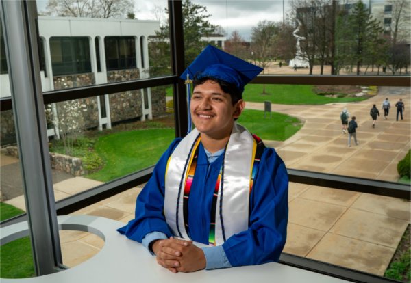 A person standing before windows and wearing an academic cap and gown smiles with hands folded before them. Buildings, art and people are in the background.