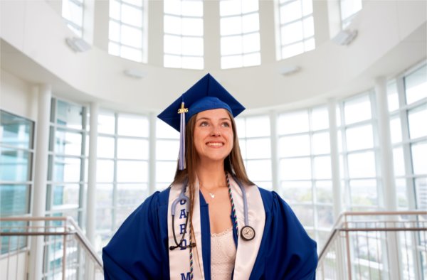 A person wearing a cap and gown stands with arms akimbo in an atrium, smiling while looking off to the side.