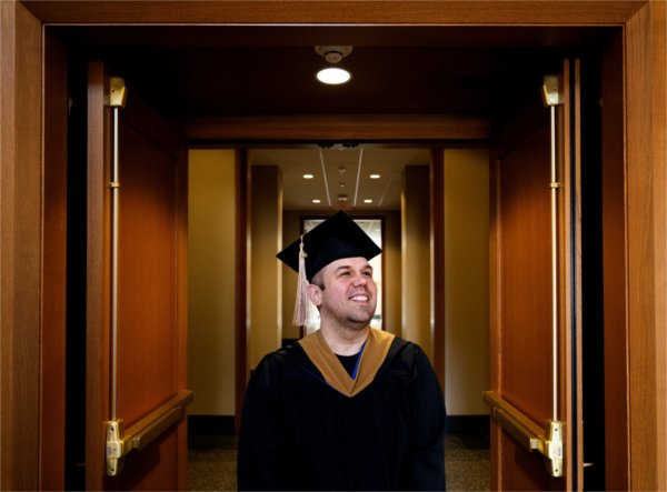A person wearing a cap and gown smiles while looking into the distance. The person is standing in a hallway.