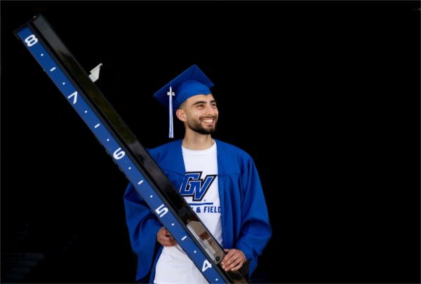 A person wearing a cap and gown and GV track and field shirt smiles while holding on to a measuring stick.