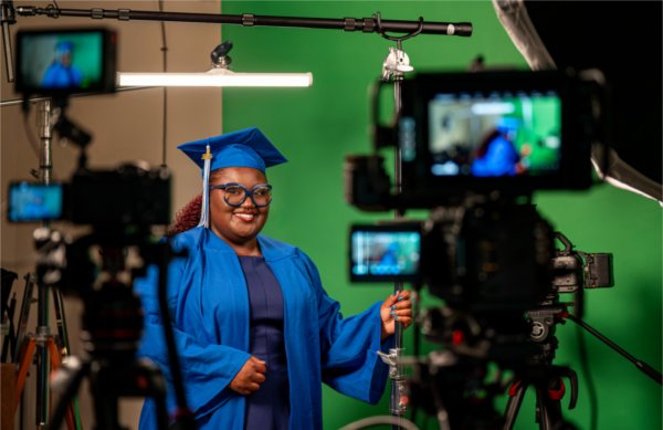A person in a cap and gown smiles while standing next to a large camera used for video production.
