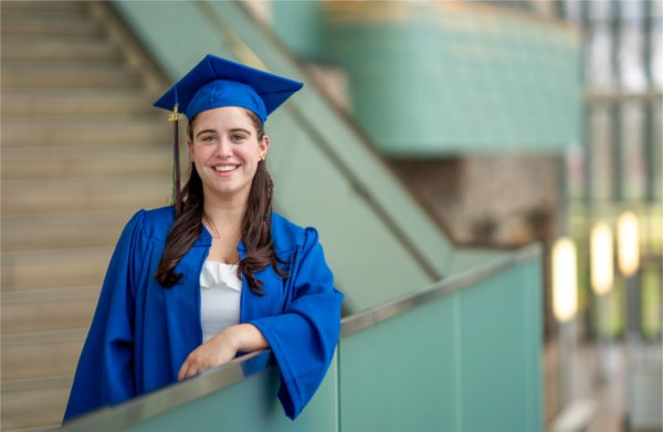 A person wearing a cap and gown smiles while leaning their arm on a railing.