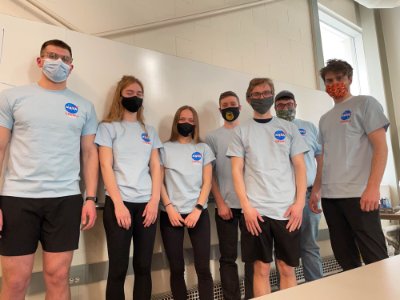 Seven students in NASA crew t-shirts standing against a wall and wearing face masks.
