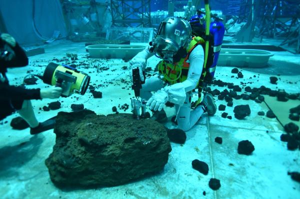Two people are underwater in a tank with big rocks on the bottom. They are in scuba suits; one has a tool to drill into a rock.