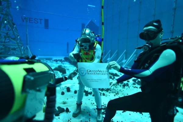 Two people in scuba suits are underwater in a tank and holding a sign that reads GV Moon Miners, with a GVSU logo
