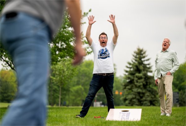  People react with arms raised during a cornhole game on a grassy lawn. 