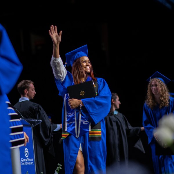 A person in a cap and gown walks across the stage with their hand up waving at the crowd.