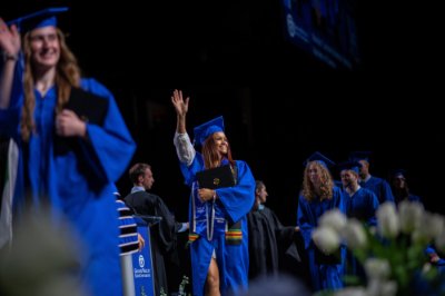 A person in a cap and gown walks across the stage with their hand up waving at the crowd. 
