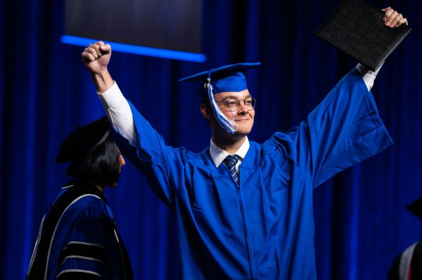 A graduate celebrates on stage after receiving their diploma with their hands up high above their head.